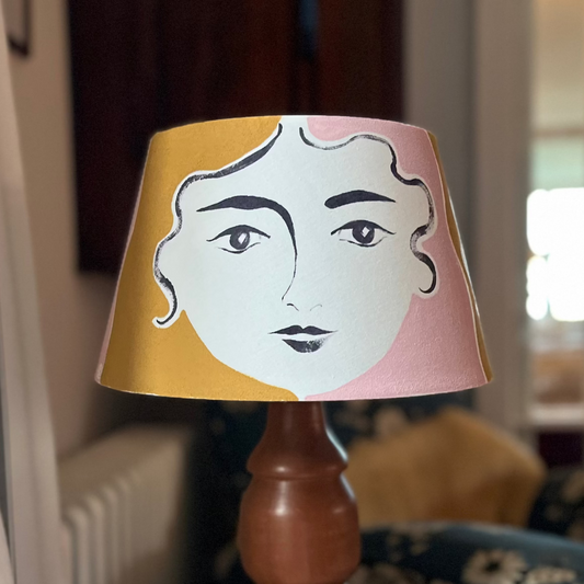 Lamp shade with hand-painted face design in Rose Pink and India Yellow sitting on vintage wooden base